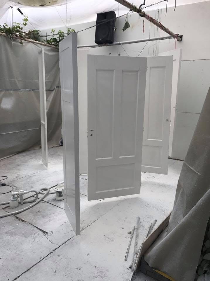 Białe, świeżo pomalowane drzwi w warsztacie, wiszące na linkach. Widoczne narzędzia do malowania natryskowego i ślady farby na podłodze.