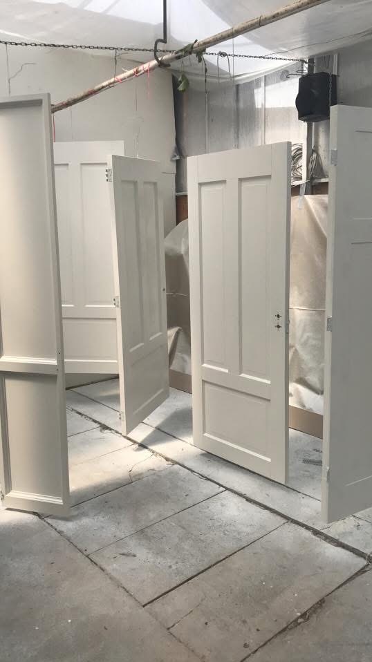 Białe, świeżo pomalowane drzwi wewnętrzne, wiszące na metalowej konstrukcji w warsztacie. Widoczne detale: zawiasy, zamek i panele. Podłoga betonowa.