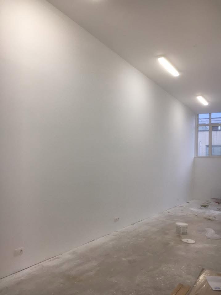 Wnętrze po malowaniu: gładkie, białe ściany, surowa podłoga, widoczne gniazdka. Minimalistyczny efekt odświeżenia z widokiem na okno w tle.