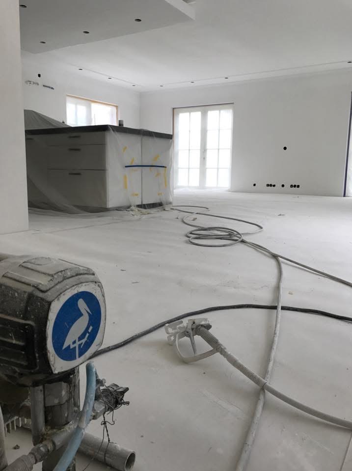 Wnętrze po malowaniu natryskowym: białe ściany, zakryta folią wyspa kuchenna, przewody i urządzenie do malowania na podłodze. Widoczne punkty świetlne w suficie.