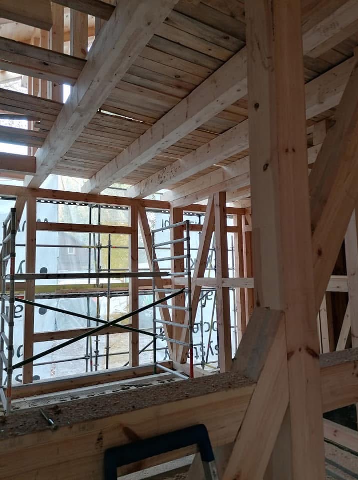 Wnętrze domu szkieletowego w trakcie budowy. Widoczne drewniane elementy konstrukcyjne, rusztowania i folia ochronna na oknach. Konstrukcja dachu z desek.