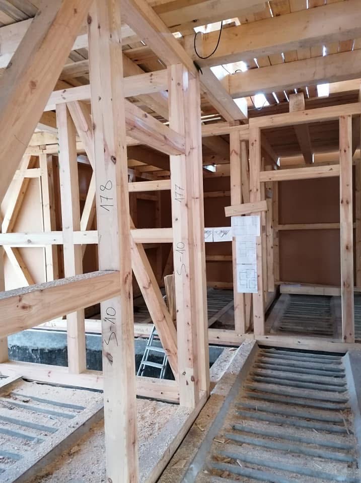 Konstrukcja domu szkieletowego w trakcie budowy. Widoczne drewniane ramy z oznaczeniami, schodki i plany architektoniczne na ścianie.