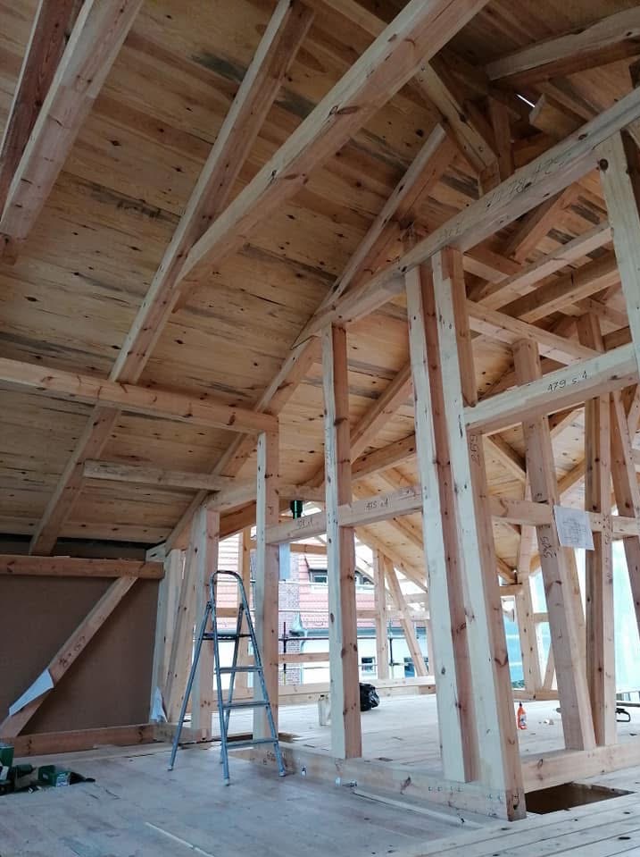 Wnętrze domu w trakcie budowy szkieletowej z widoczną drewnianą konstrukcją dachu, ścian i podłogi. Na podłodze stoi drabina, a w tle widać budynki.