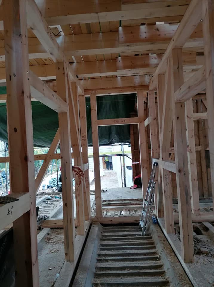 Wnętrze domu w trakcie budowy, widoczna drewniana konstrukcja szkieletowa, schody betonowe i drabina oparta o ścianę. Surowy stan, widoczne elementy instalacji.