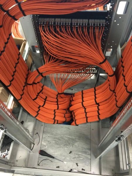 Starannie ułożone pomarańczowe kable w szafie rack, spięte czarnymi opaskami. Widoczne złącza i elementy montażowe. Narzędzie leży na podłodze szafy.