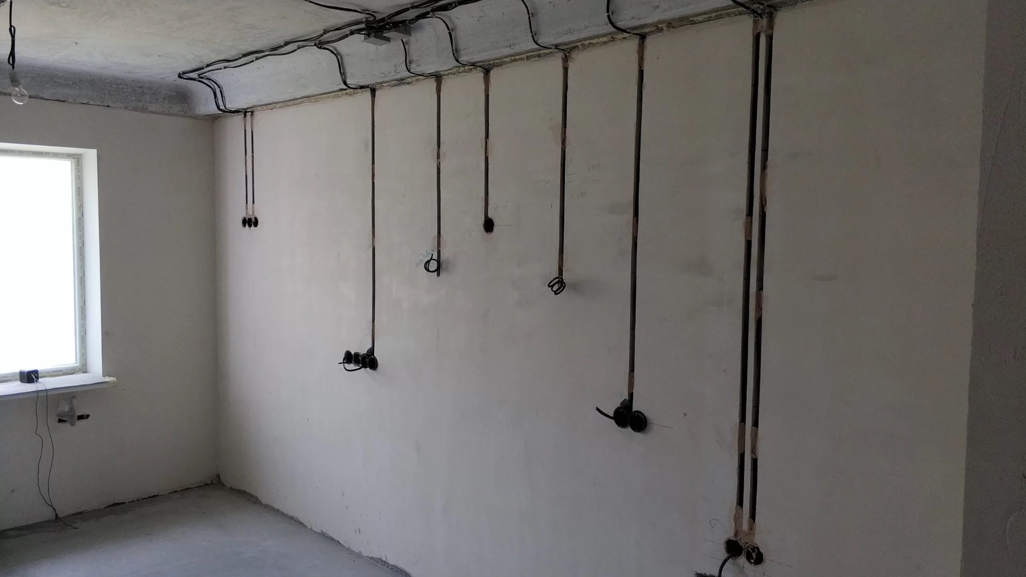 Surowy pokój z widoczną instalacją elektryczną: przewody biegnące po ścianach i suficie, przygotowane punkty do gniazdek i oświetlenia, okno z parapetem.