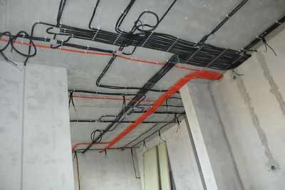 Surowe wnętrze w trakcie prac elektrycznych: widoczne czarne i pomarańczowe kable prowadzone po suficie i ścianach, przytwierdzone uchwytami, surowy beton.