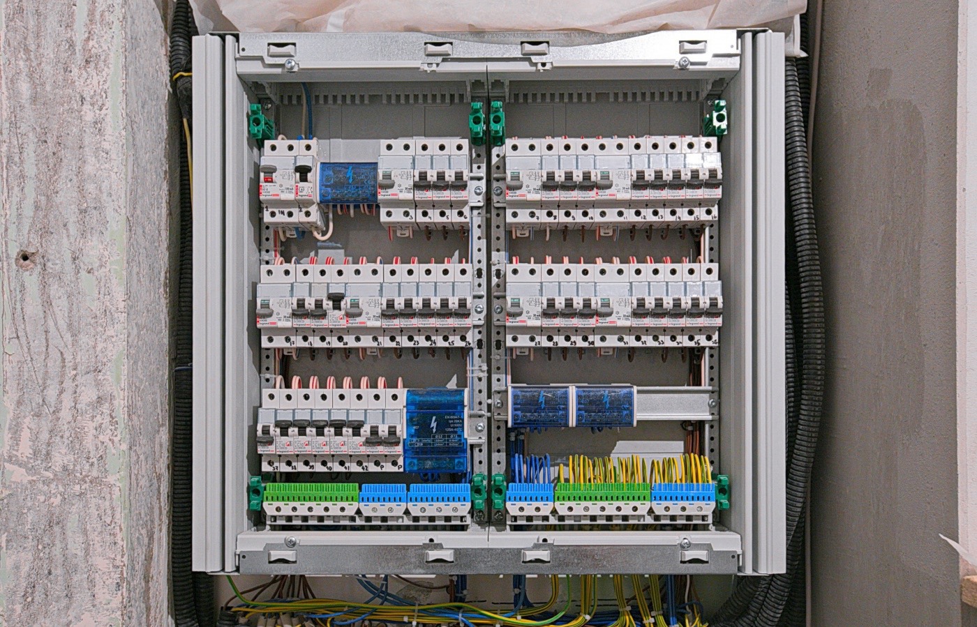 Rozdzielnica elektryczna z licznymi zabezpieczeniami, zamontowana na ścianie w surowym stanie. Widoczne kable w peszlach i liczne przewody.