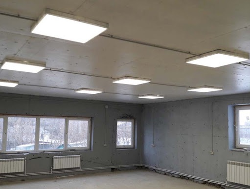 Nowoczesne, kwadratowe panele LED równomiernie rozmieszczone na suficie surowego, szarego pomieszczenia z oknami i grzejnikami. Widoczne przewody elektryczne.
