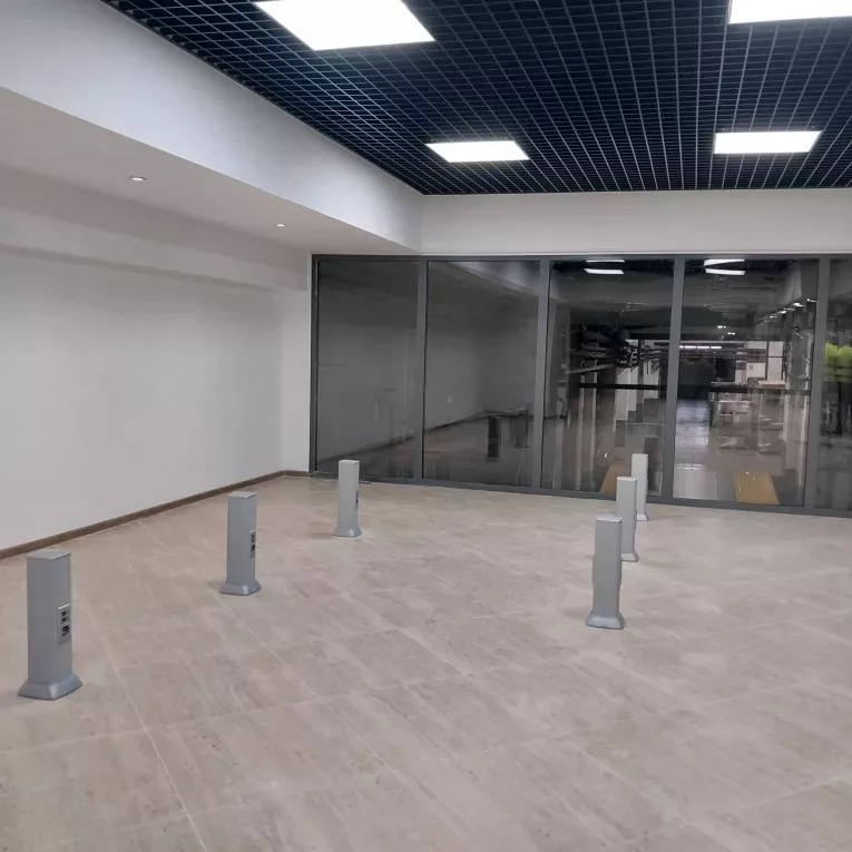 Wnętrze z szarą podłogą, szklaną ścianą i rzędami słupków z gniazdkami elektrycznymi. Sufit kasetonowy z oświetleniem.