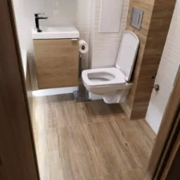 Nowoczesna łazienka po remoncie: wisząca toaleta, umywalka z szafką w odcieniach drewna, płytki podłogowe imitujące drewno, minimalistyczne dodatki.