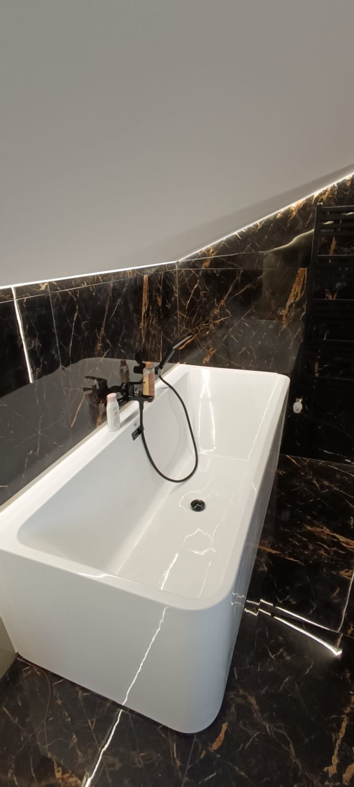 Nowoczesna, biała wanna na tle czarnej marmurowej ściany z delikatnym oświetleniem LED. Widoczne baterie i akcesoria łazienkowe.