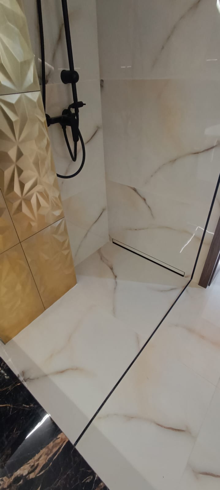 Wykończona kabina prysznicowa z czarną armaturą i odpływem liniowym. Ściany wyłożone płytkami imitującymi marmur, jedna ściana z dekoracyjną, złotą płytką 3D.