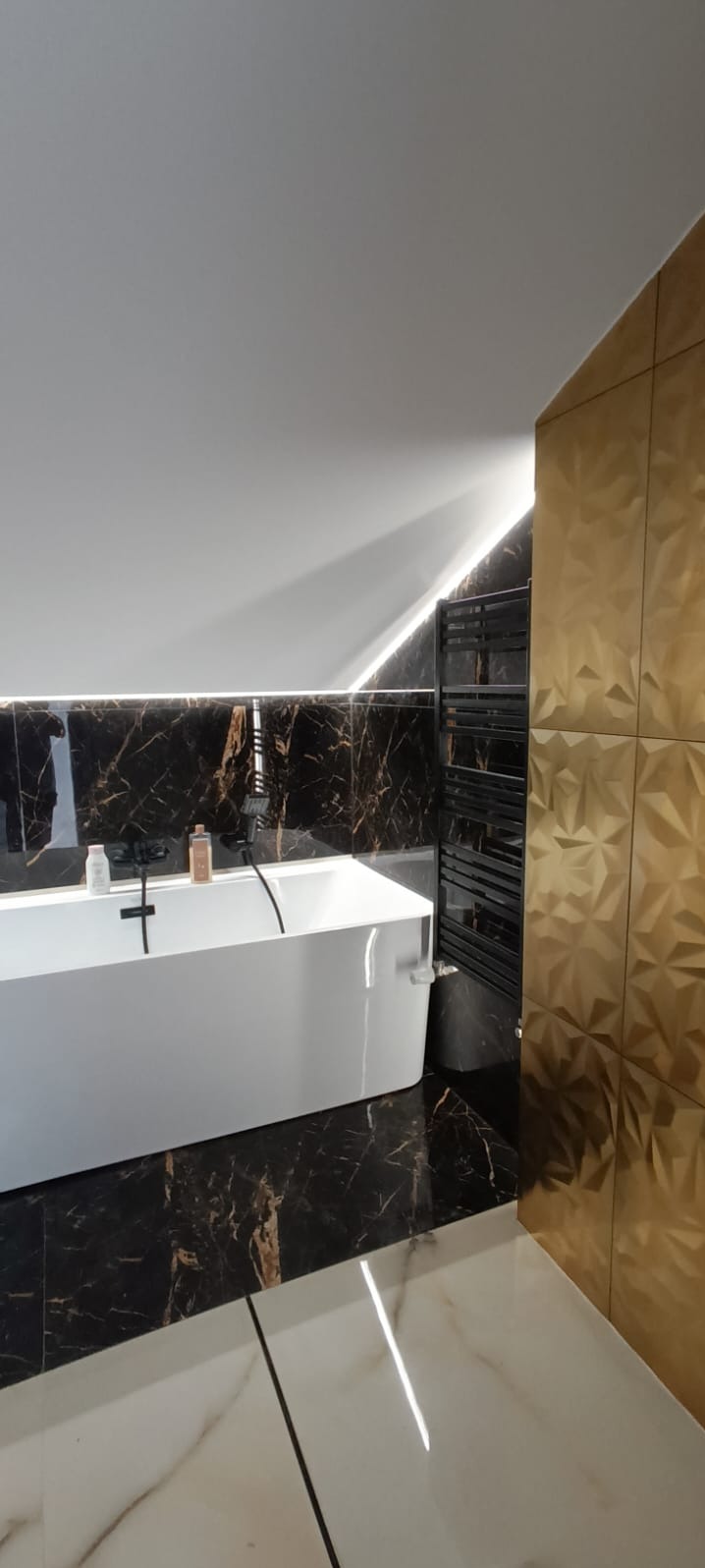 Nowoczesna łazienka na poddaszu z czarno-złotymi płytkami imitującymi marmur, białą wanną i grzejnikiem drabinkowym. Uwagę przykuwają złote, geometryczne płytki ścienne.