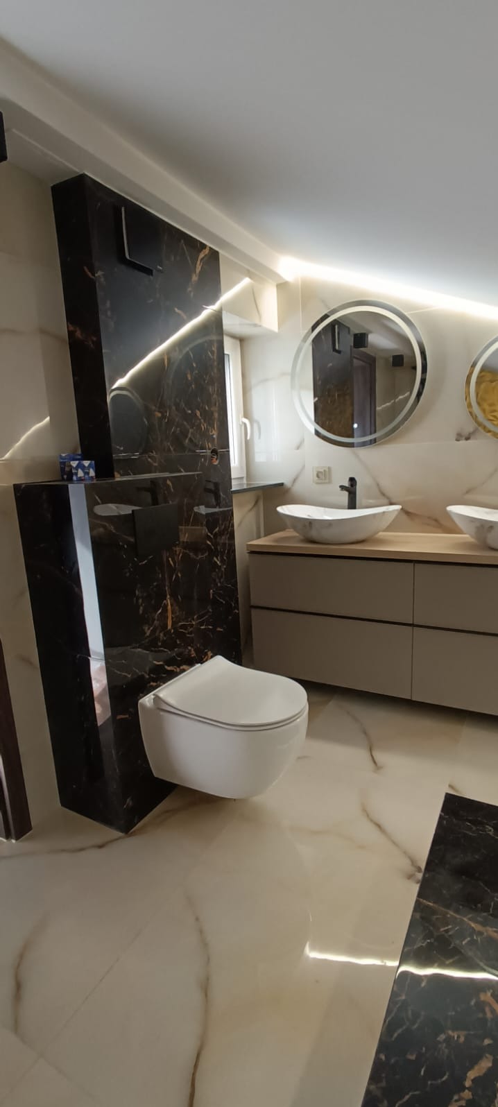 Elegancka łazienka w beżach i czerni: podwieszana toaleta, umywalki na szafce z okrągłymi lustrami z podświetleniem LED, marmurowe płytki na podłodze i ścianach.