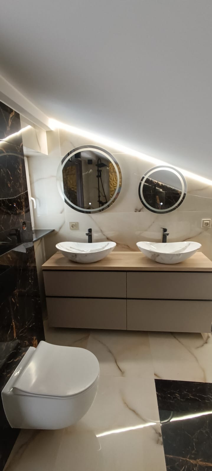 Nowoczesna łazienka z dwoma okrągłymi lustrami z podświetleniem LED, marmurowymi umywalkami na drewnianej szafce i podwieszaną toaletą. Czarne i białe płytki marmurowe.