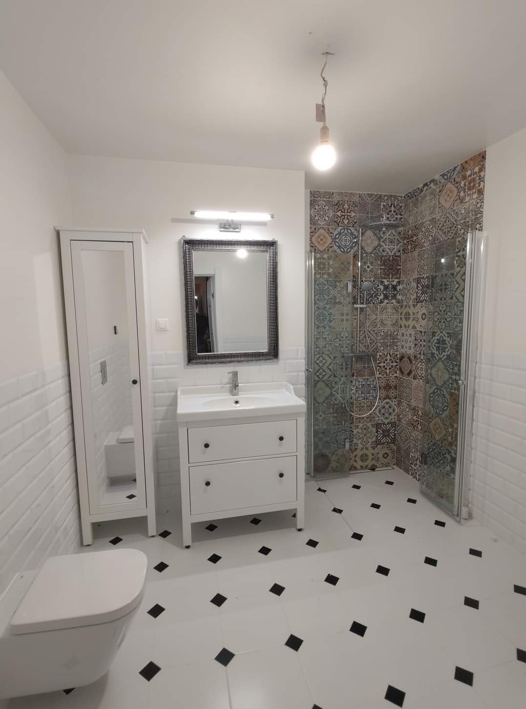 Wykończona łazienka z białą armaturą, czarno-białą podłogą i kabiną prysznicową z marokańskimi wzorami na płytkach. Widoczna toaleta, szafka z umywalką i lustro.