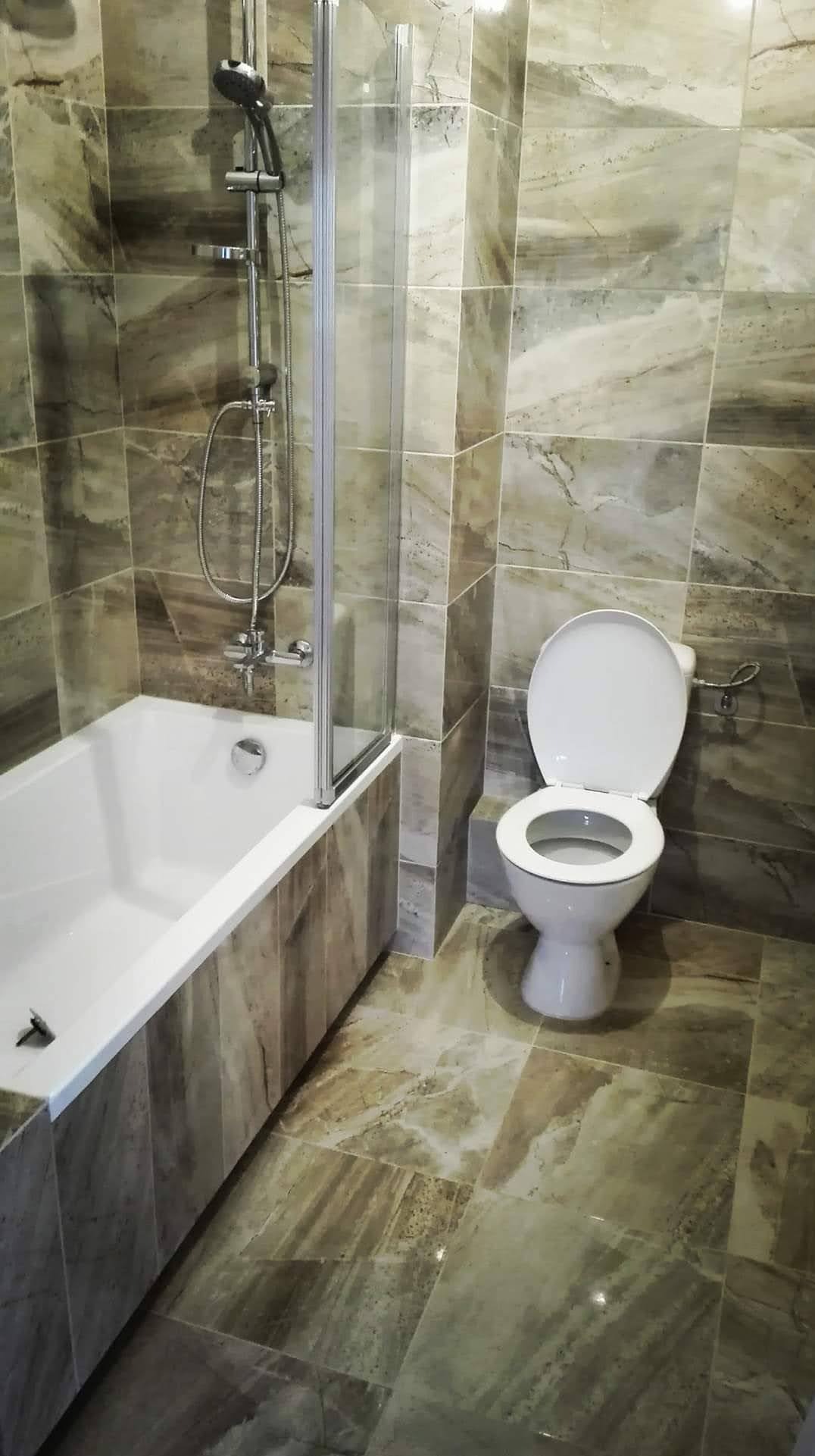 Wykończona łazienka w beżowo-brązowej tonacji z wanną z parawanem, toaletą i płytkami imitującymi marmur na ścianach i podłodze. Widok na całe pomieszczenie.