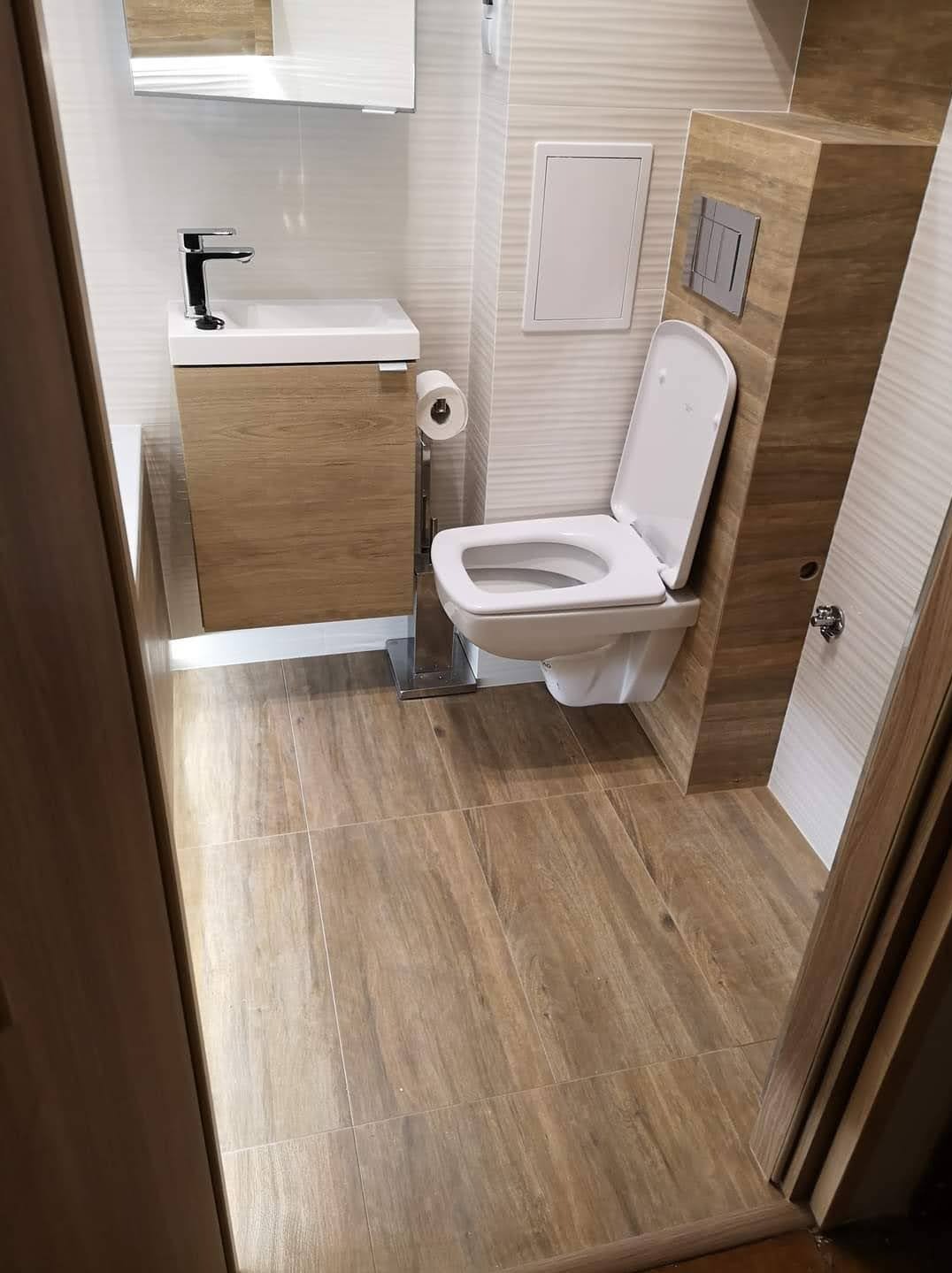 Nowoczesna łazienka po remoncie: wisząca toaleta, umywalka z szafką w odcieniach drewna, płytki podłogowe imitujące drewno, minimalistyczne dodatki.