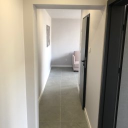 Bogacz-Studziński - Wykończony korytarz w neutralnych barwach z szarą podłogą z płytek i widokiem na pokój z jasnoróżowym fotelem. Minimalistyczny design, nowoczesne wykończenie.