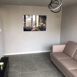 Bogacz-Studziński - Salon we Wrocławiu po odświeżeniu: szare ściany, grafitowa podłoga z płytek, nowoczesna lampa wisząca i abstrakcyjny obraz nad sofą.