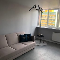 Bogacz-Studziński - Jasny salon z szarą ścianą, beżową sofą z poduszkami w kolorze butelkowej zieleni, nowoczesną lampą i widokiem na blok przez okno.