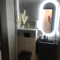 Nowoczesna, szara łazienka z podświetlanym lustrem, czarną armaturą i dekoracyjnymi trawami pampasowymi. Minimalistyczny design, płytki imitujące kamień.