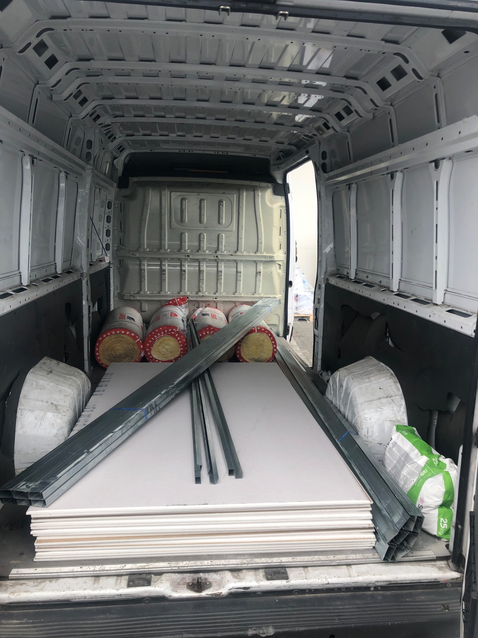 Wnętrze białego busa z materiałami budowlanymi: płyty karton-gips, profile metalowe, wełna mineralna w rolkach i worek zaprawy. Widok z tyłu pojazdu.
