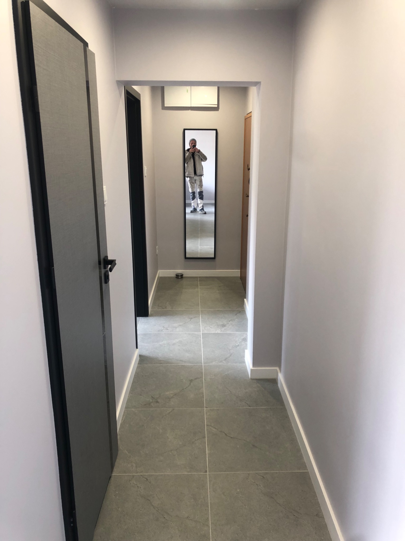 Wykończony korytarz w szarościach z lustrem, w którym odbija się osoba robiąca zdjęcie. Nowe płytki na podłodze, świeżo pomalowane ściany i nowe drzwi.