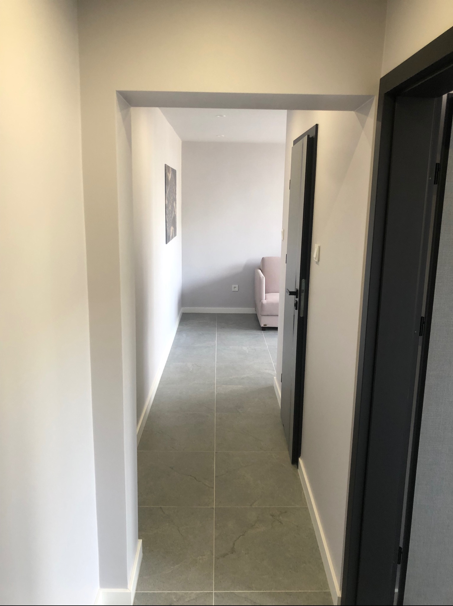 Wykończony korytarz w neutralnych barwach z szarą podłogą z płytek i widokiem na pokój z jasnoróżowym fotelem. Minimalistyczny design, nowoczesne wykończenie.
