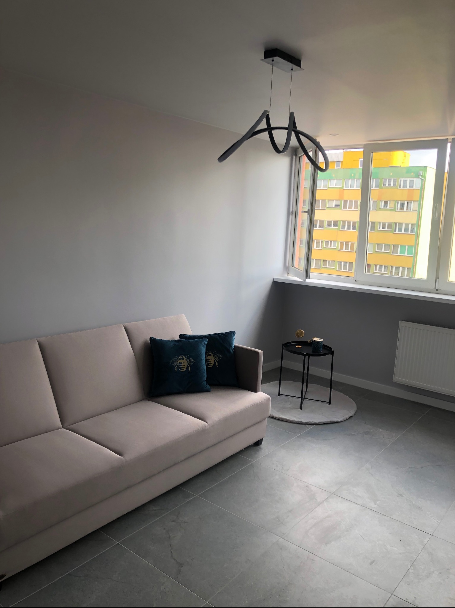 Jasny salon z szarą ścianą, beżową sofą z poduszkami w kolorze butelkowej zieleni, nowoczesną lampą i widokiem na blok przez okno.