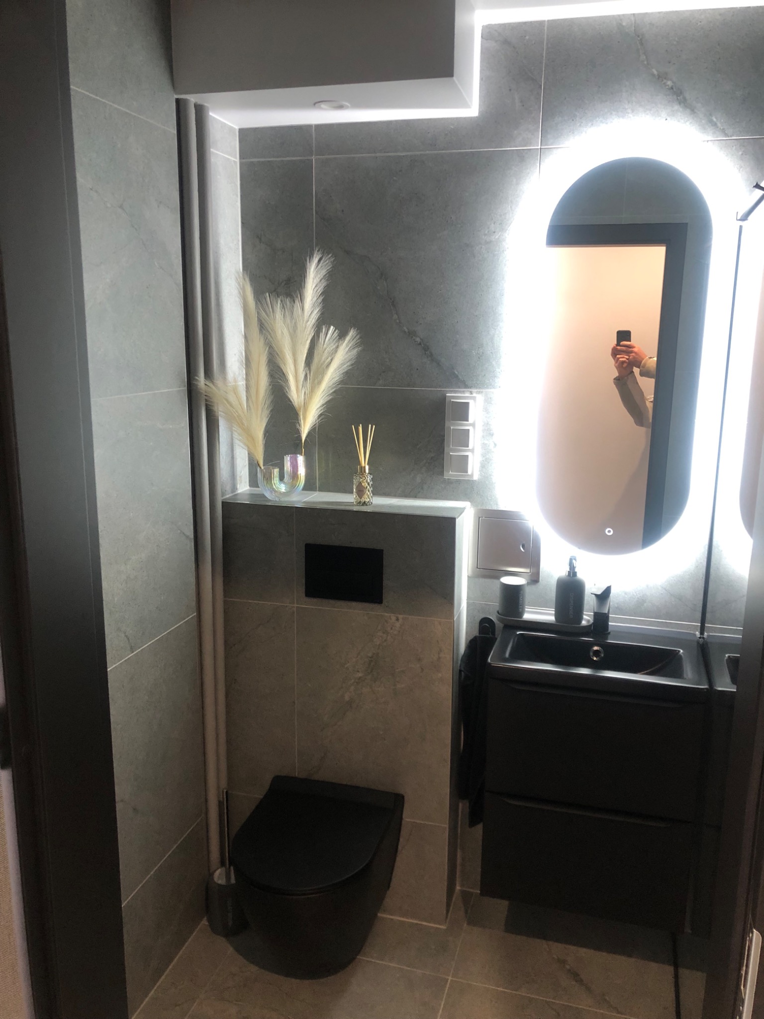 Nowoczesna, szara łazienka z podświetlanym lustrem, czarną armaturą i dekoracyjnymi trawami pampasowymi. Minimalistyczny design, płytki imitujące kamień.