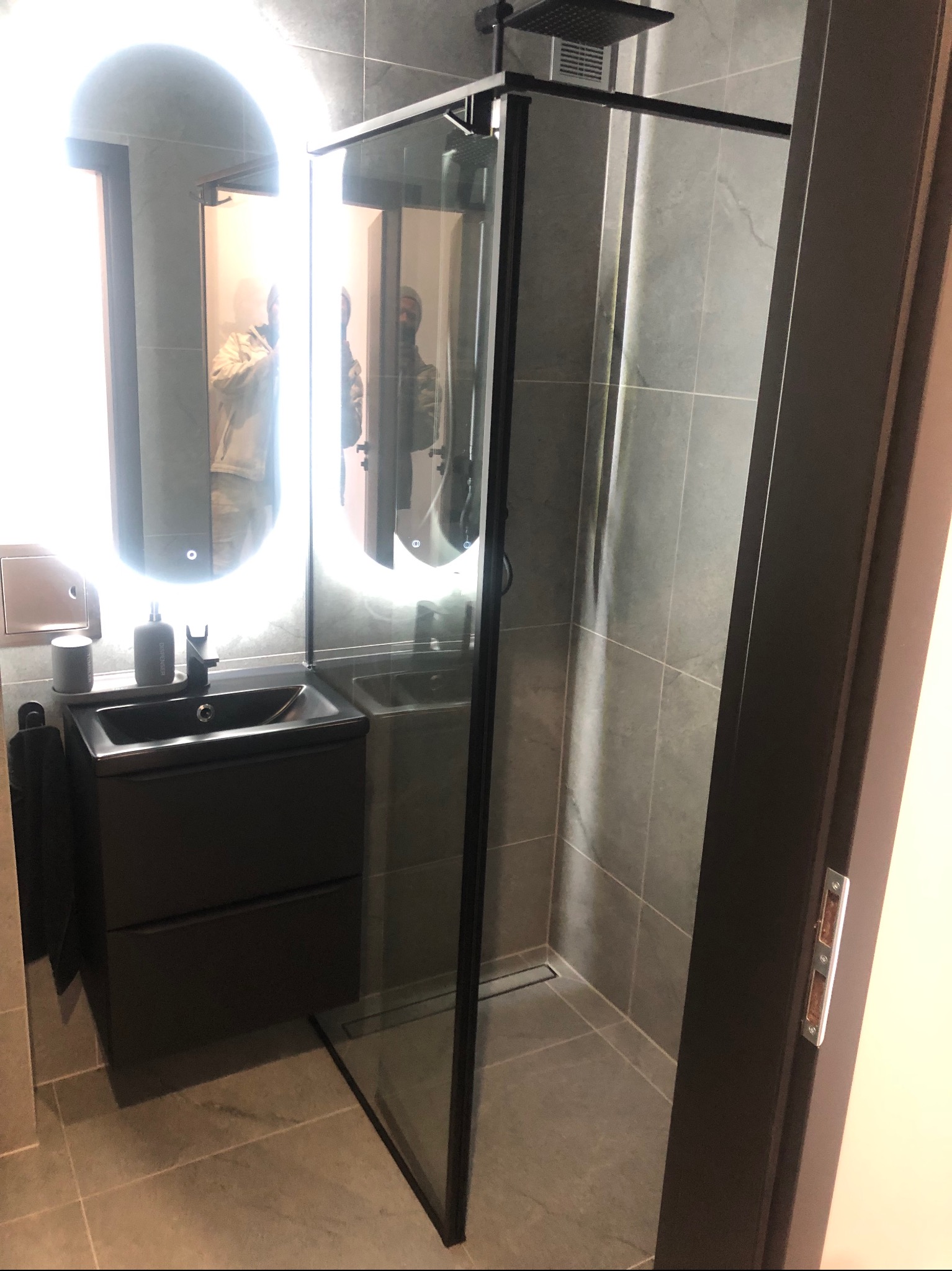 Nowoczesna łazienka z kabiną prysznicową typu walk-in, umywalką w kolorze antracytu i podświetlanym lustrem. Minimalistyczny design z szarymi płytkami.