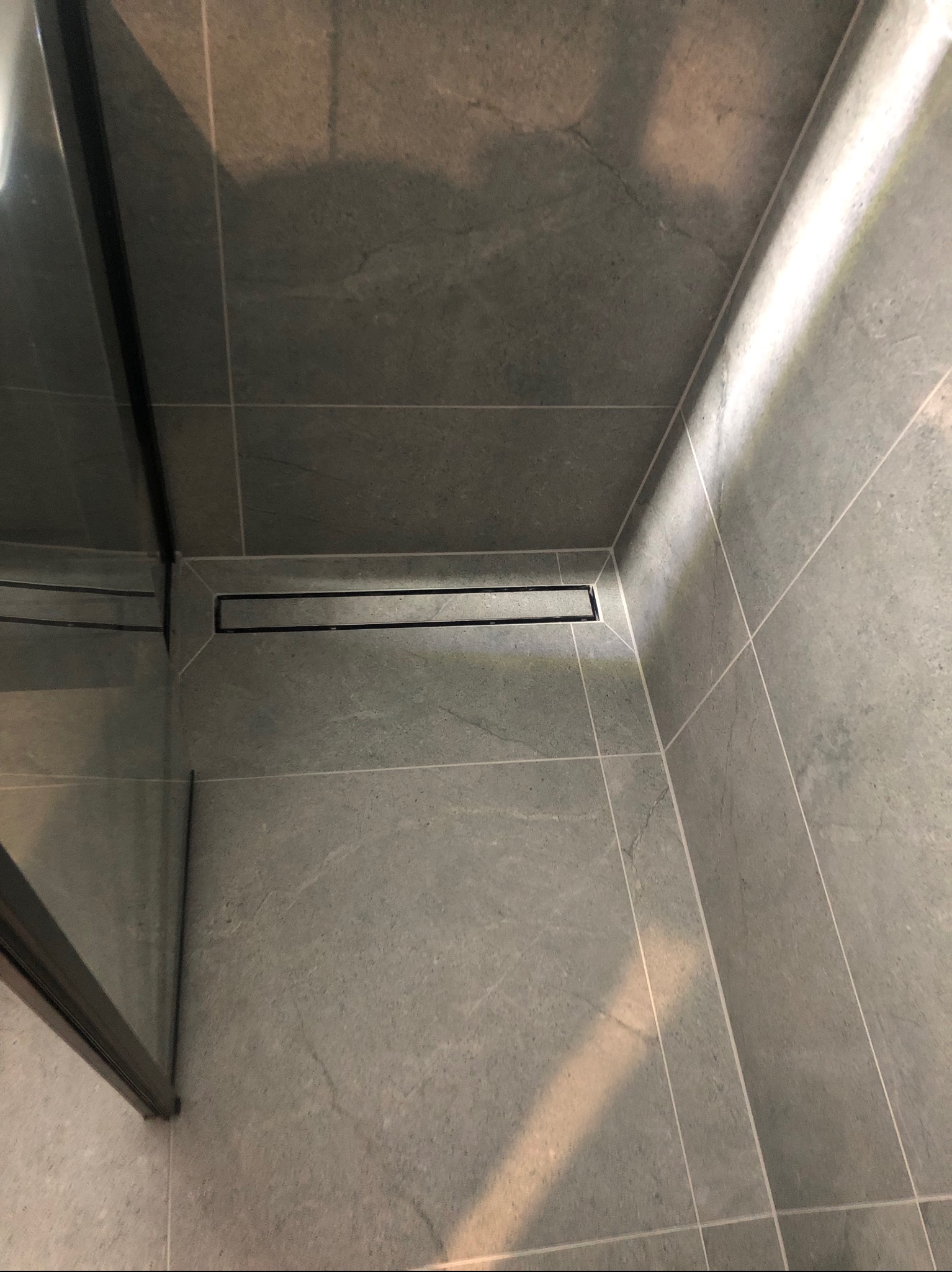Wykończona kabina prysznicowa z szarymi płytkami i odpływem liniowym. Widoczne fugi i fragment szklanej ścianki.