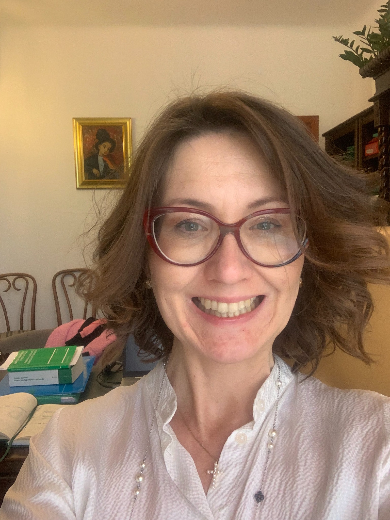 Uśmiechnięta brunetka w okularach, z książkami i obrazem w tle. Portret kobiety w białej koszuli z naszyjnikiem, siedzącej przy biurku w domowym biurze.