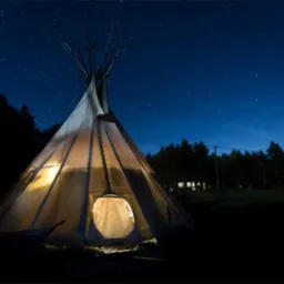 Tipi oświetlone od wewnątrz na tle nocnego, rozgwieżdżonego nieba. W tle widoczny las i budynek z oknami.