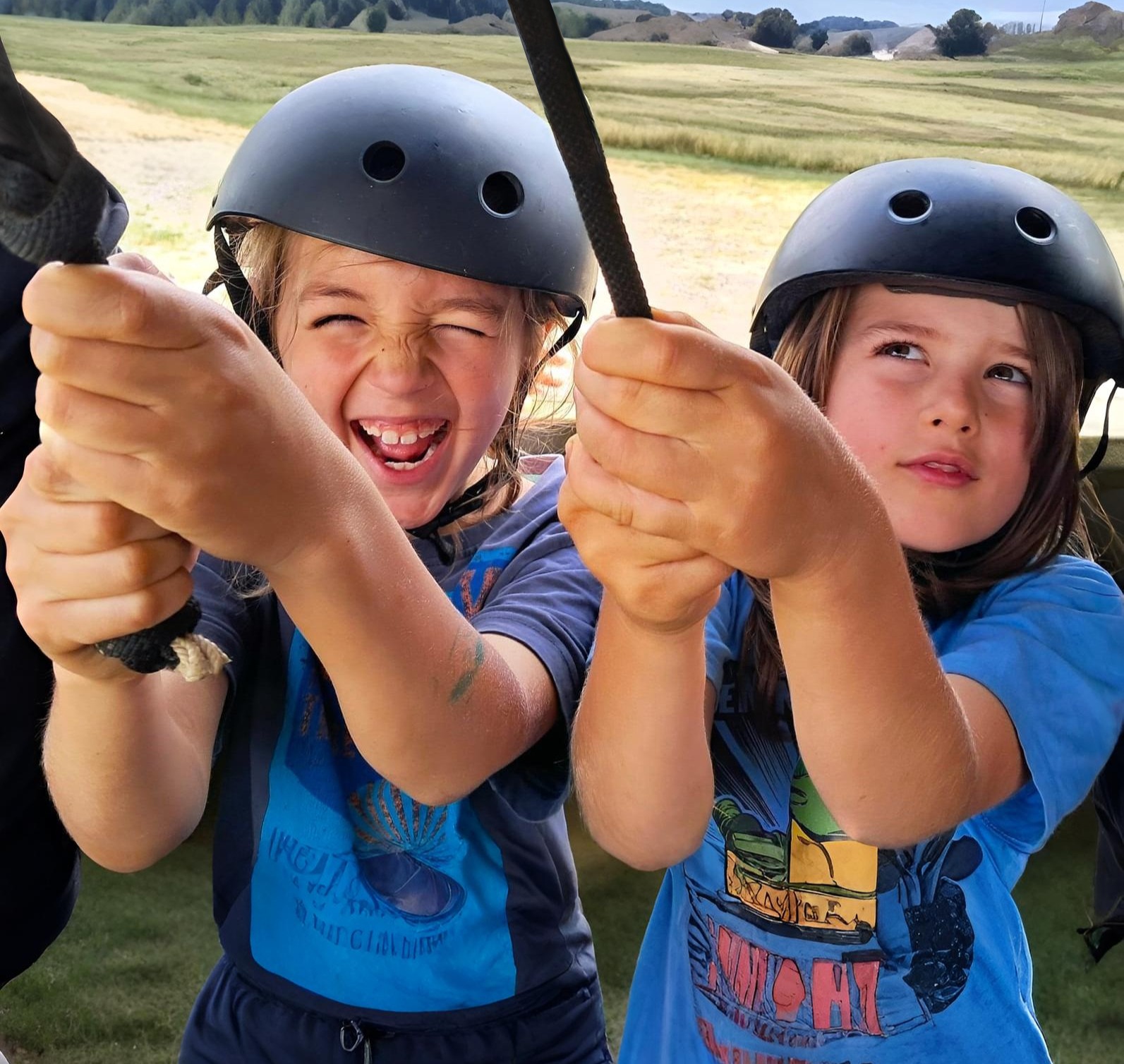 Dwoje dzieci w kaskach, trzymających linę na tle zielonego krajobrazu. Jedno dziecko roześmiane, drugie patrzy w górę. Aktywność na świeżym powietrzu.