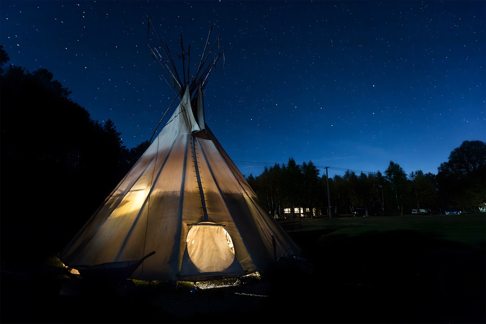 Tipi oświetlone od wewnątrz na tle nocnego, rozgwieżdżonego nieba. W tle widoczny las i budynek z oknami.