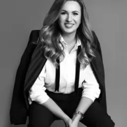 Portret kobiety w czarno-białej tonacji. Uśmiechnięta, w eleganckiej koszuli i spodniach, z marynarką na ramieniu. Minimalistyczne tło, stonowana kolorystyka.