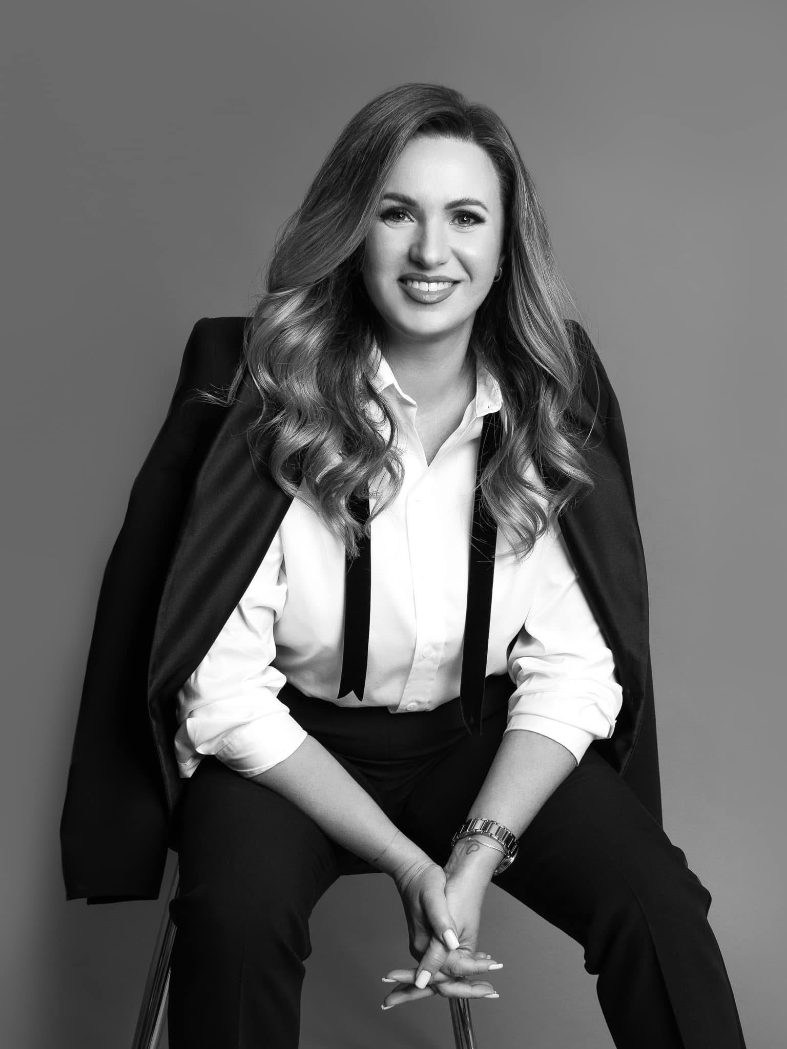 Portret kobiety w czarno-białej tonacji. Uśmiechnięta, w eleganckiej koszuli i spodniach, z marynarką na ramieniu. Minimalistyczne tło, stonowana kolorystyka.