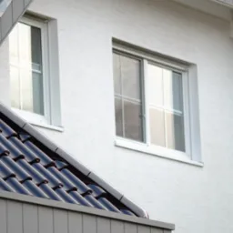 Fragment elewacji domu z oknami i błyszczącą, czarną dachówką. Widoczna faktura tynku i precyzyjne wykończenie detali.