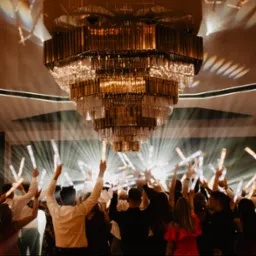 Rozświetlony parkiet pod kryształowym żyrandolem. Grupa osób z podniesionymi rękami trzymających świecące pałeczki, zabawa w trakcie przyjęcia.