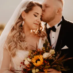 Panna młoda w koronkowej sukni ślubnej i welonie trzyma bukiet z słonecznikiem, gdy pan młody w smokingu czule całuje ją w skroń na tle gór.