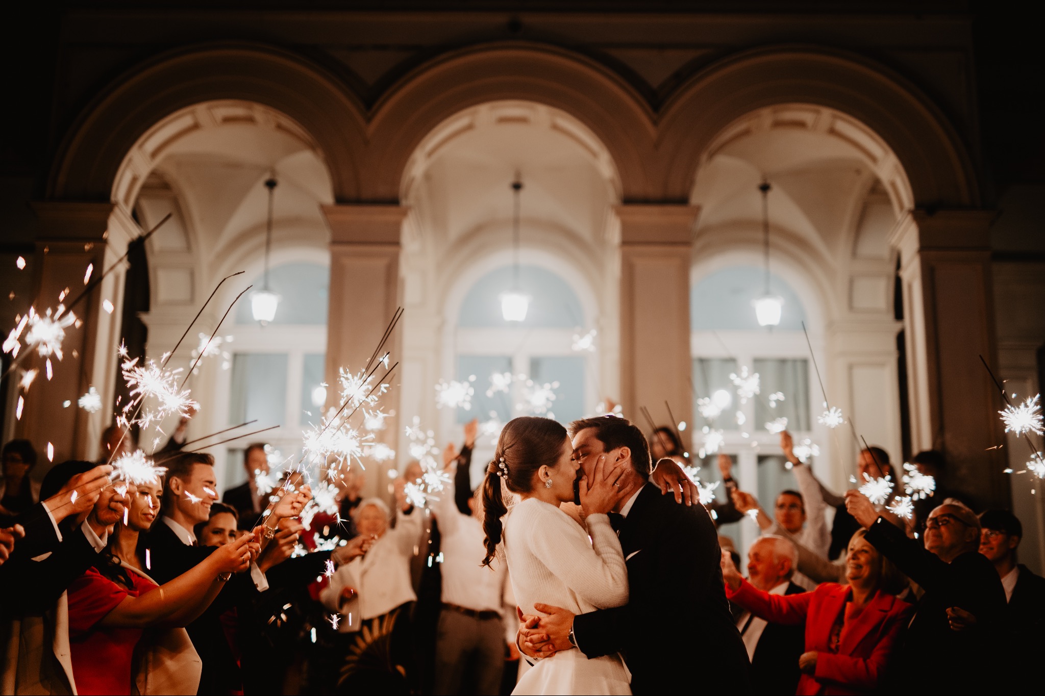 Pocałunek nowożeńców w otoczeniu gości z zimnymi ogniami na tle architektury z łukami i ciepłym oświetleniem. Ujęcie z bliska, dominują ciepłe barwy.