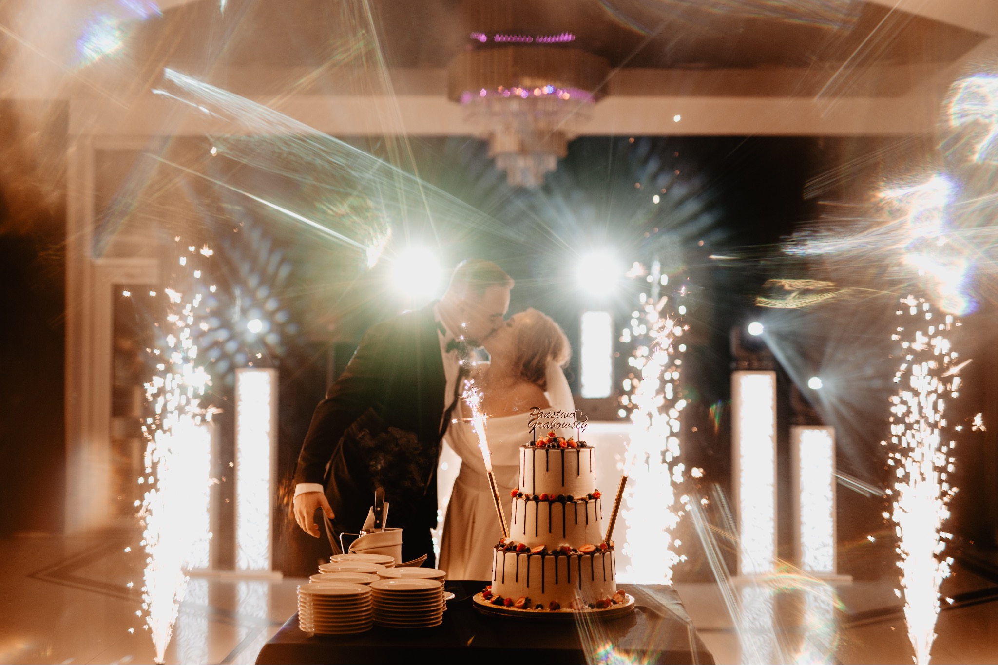 Pocałunek pary młodej za tortem weselnym, w tle fontanna iskier i dekoracje sali. Na torcie napis 'Państwo Grażowscy'.