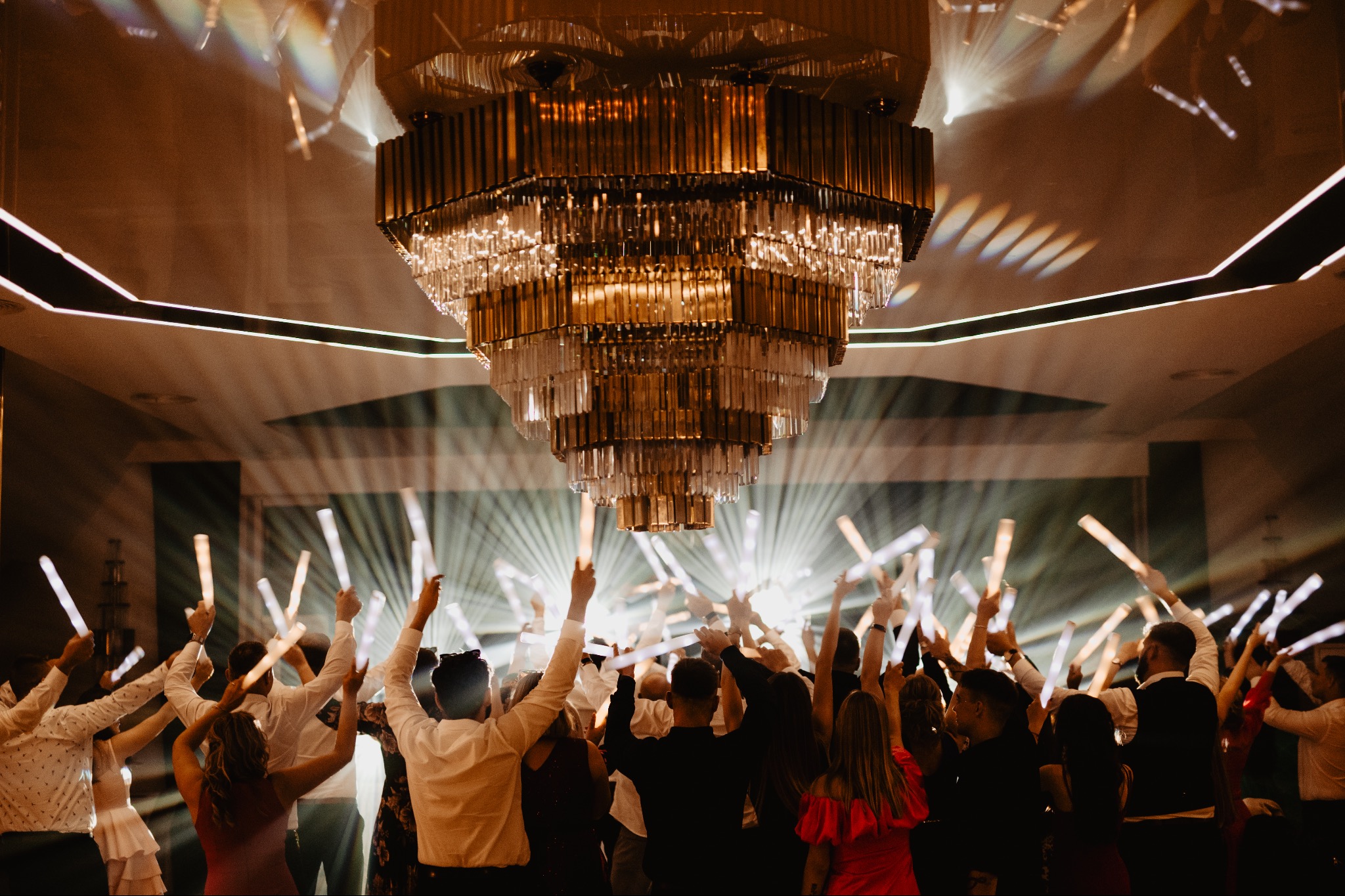 Rozświetlony parkiet pod kryształowym żyrandolem. Grupa osób z podniesionymi rękami trzymających świecące pałeczki, zabawa w trakcie przyjęcia.