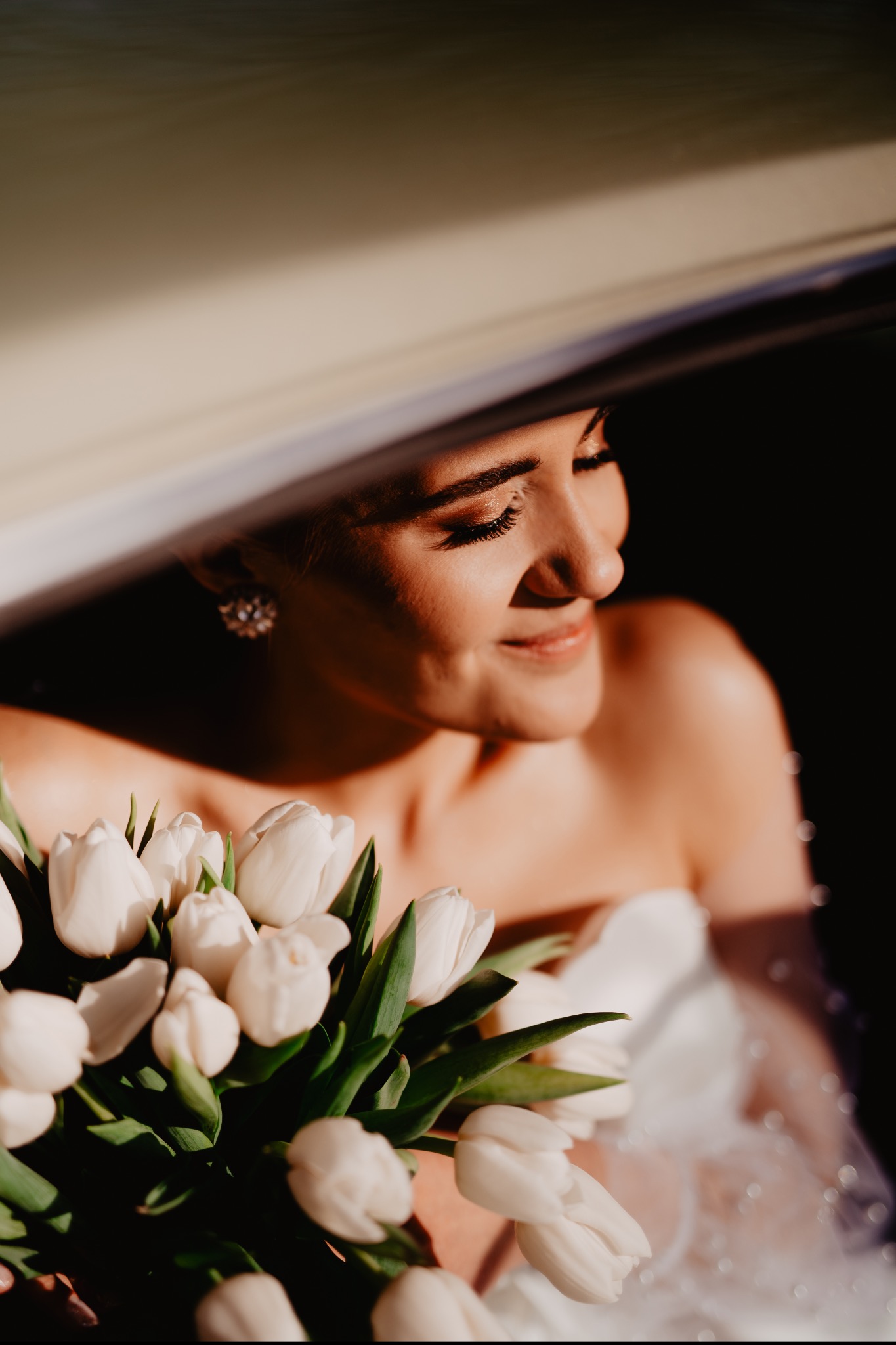 Panna młoda w samochodzie, z bukietem białych tulipanów, spogląda w bok, oświetlona ciepłym światłem. Widoczny fragment sukni i kolczyk.