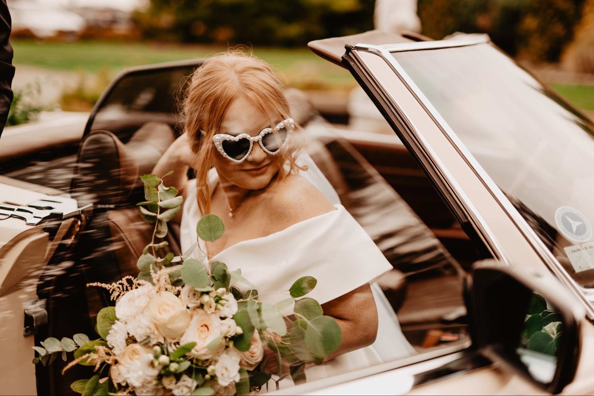 Panna młoda w białej sukni i okularach w kształcie serc siedzi w kabriolecie z bukietem róż i eukaliptusa. Mercedes z otwartym dachem w tle.