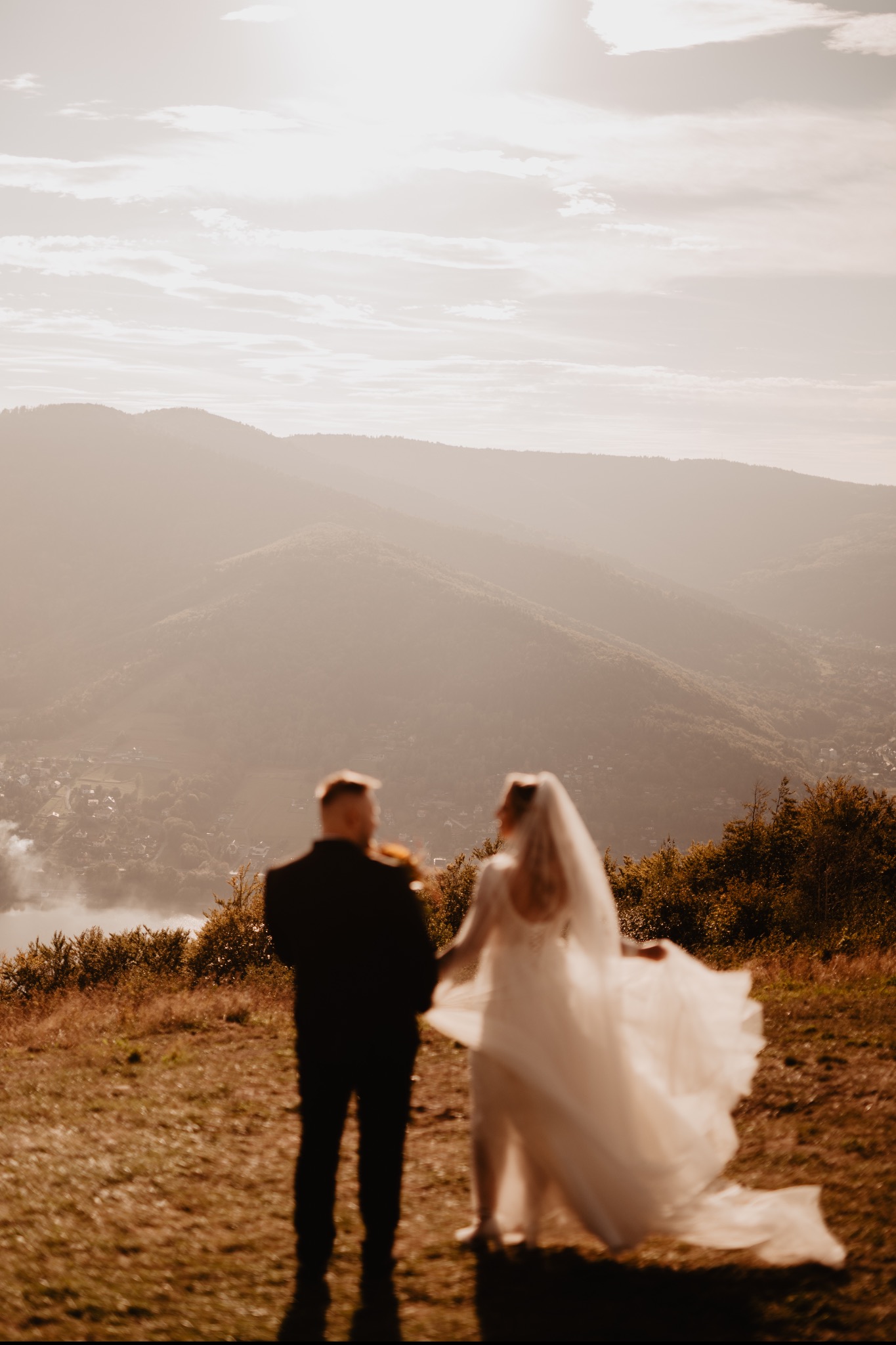 Para młoda, w sukni i garniturze, stoi tyłem na wzgórzu z widokiem na góry w lekkiej mgle. Ujęcie z oddali, z rozmytym pierwszym planem.
