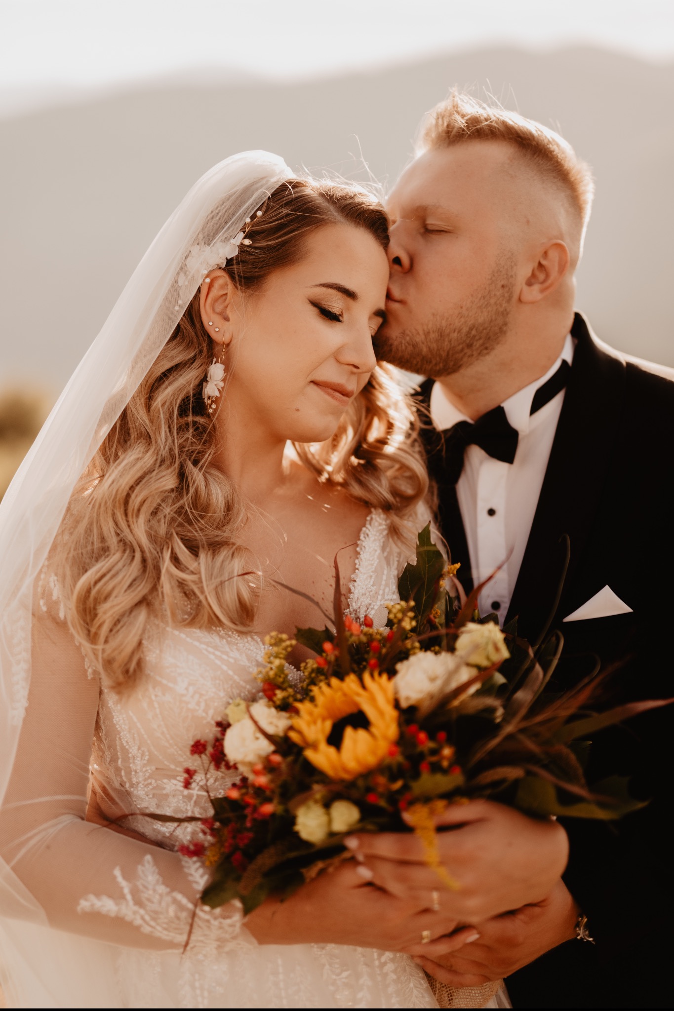 Panna młoda w koronkowej sukni ślubnej i welonie trzyma bukiet z słonecznikiem, gdy pan młody w smokingu czule całuje ją w skroń na tle gór.